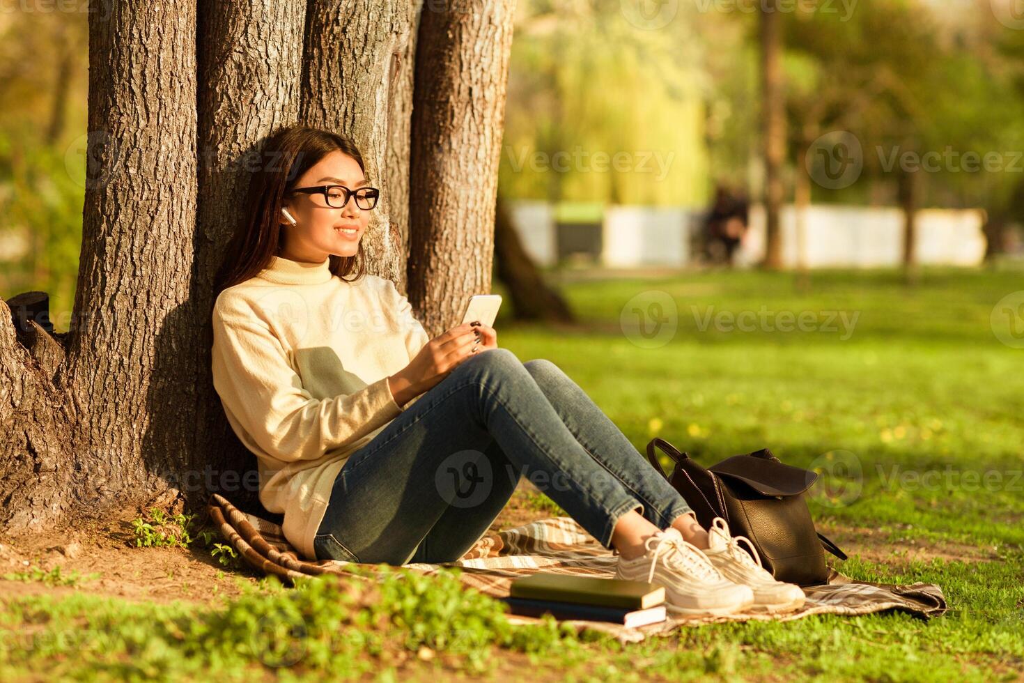 Asian young woman is relaxing under a tree in a sunny park, looking at her smartphone. She is wearing casual clothes and glasses, sitting comfortably on a blanket with books beside her photo