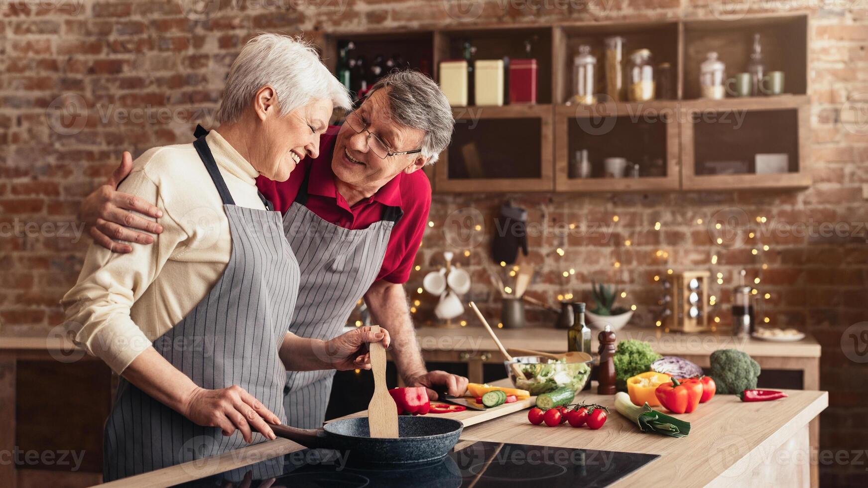 A senior couple is cooking together in a modern kitchen. The woman is stirring something in a pan on the stove while the man stands behind her, smiling and hugging her from behind. photo