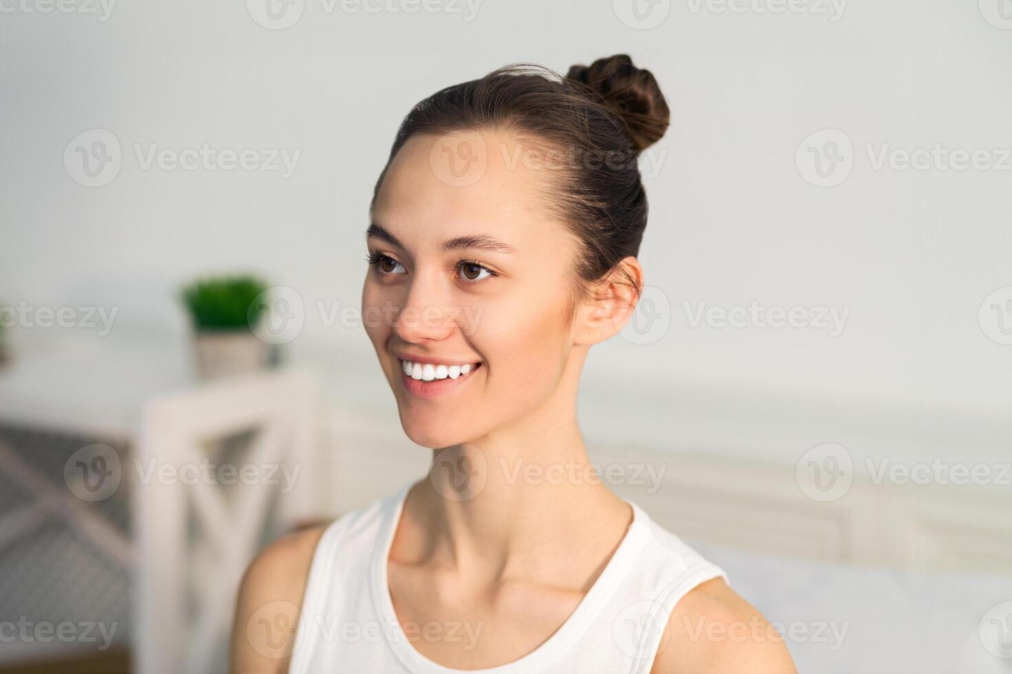 A woman with a bun smiles at the camera while sitting in a bright room. photo