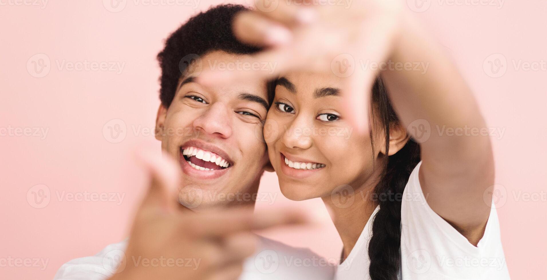 A cheerful young African American couple smiles and poses playfully with a bright pink background. They both wear white shirts and appear excited and happy, sharing a joyous moment together. photo