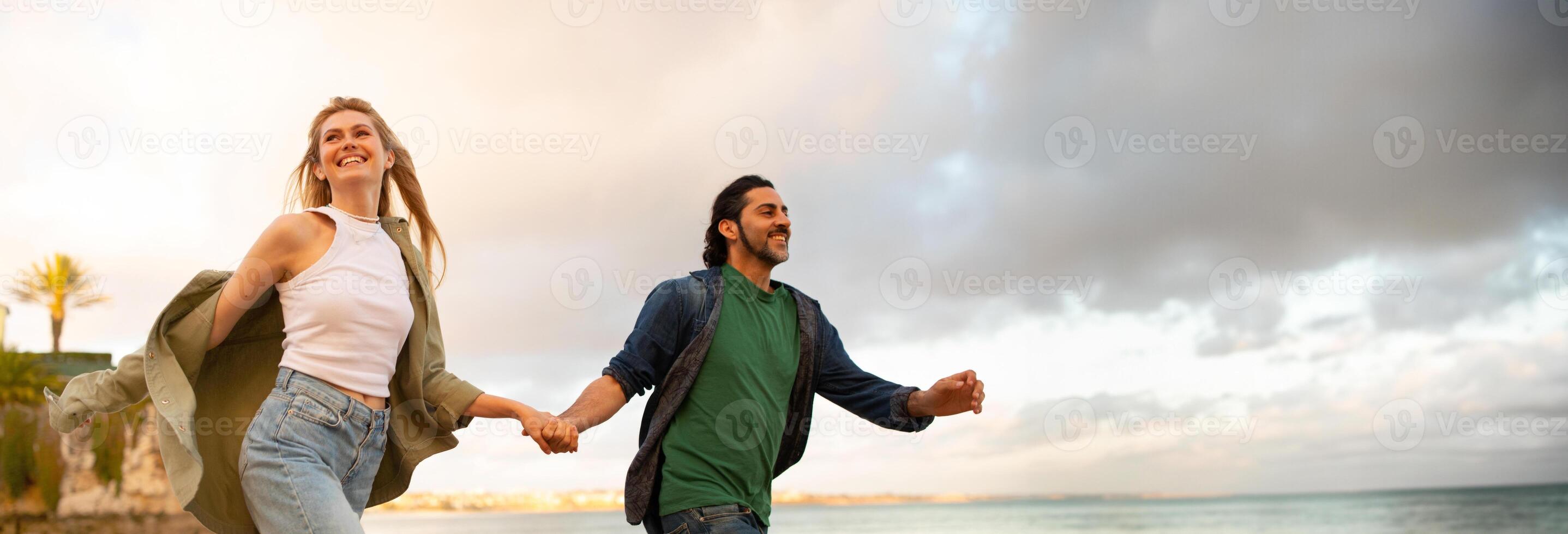 A young couple walks hand-in-hand along a beach at sunset. They are both smiling and seem to be enjoying their time together, panorama with copy space photo