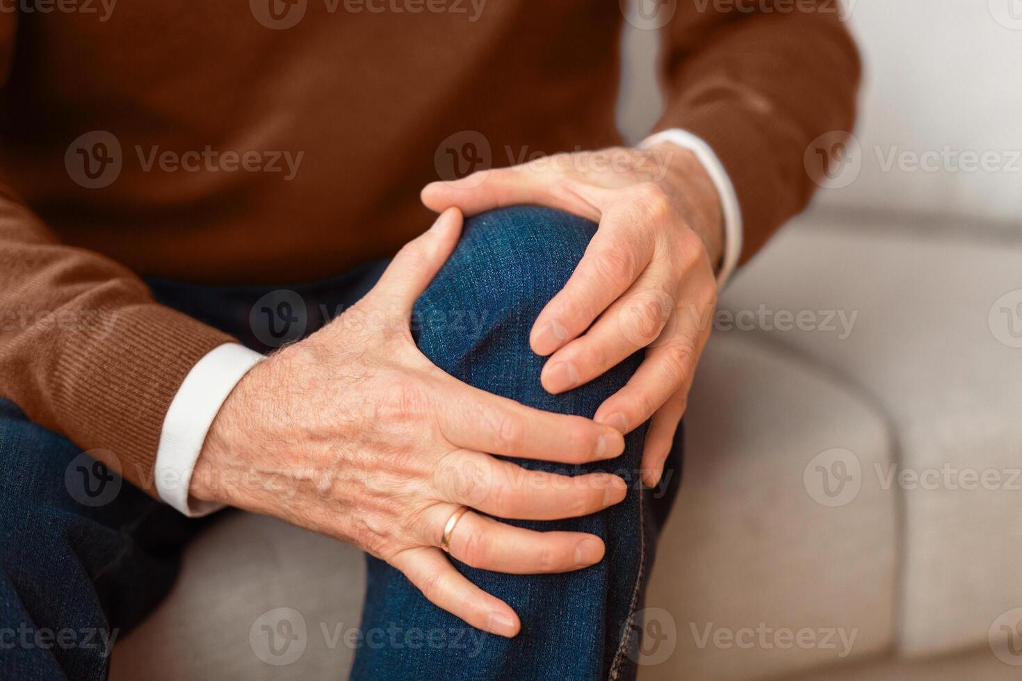 Arthritis concept. Unrecognizable elderly man massaging his knee suffering from pain sitting on couch at home. Selective focus, cropped photo