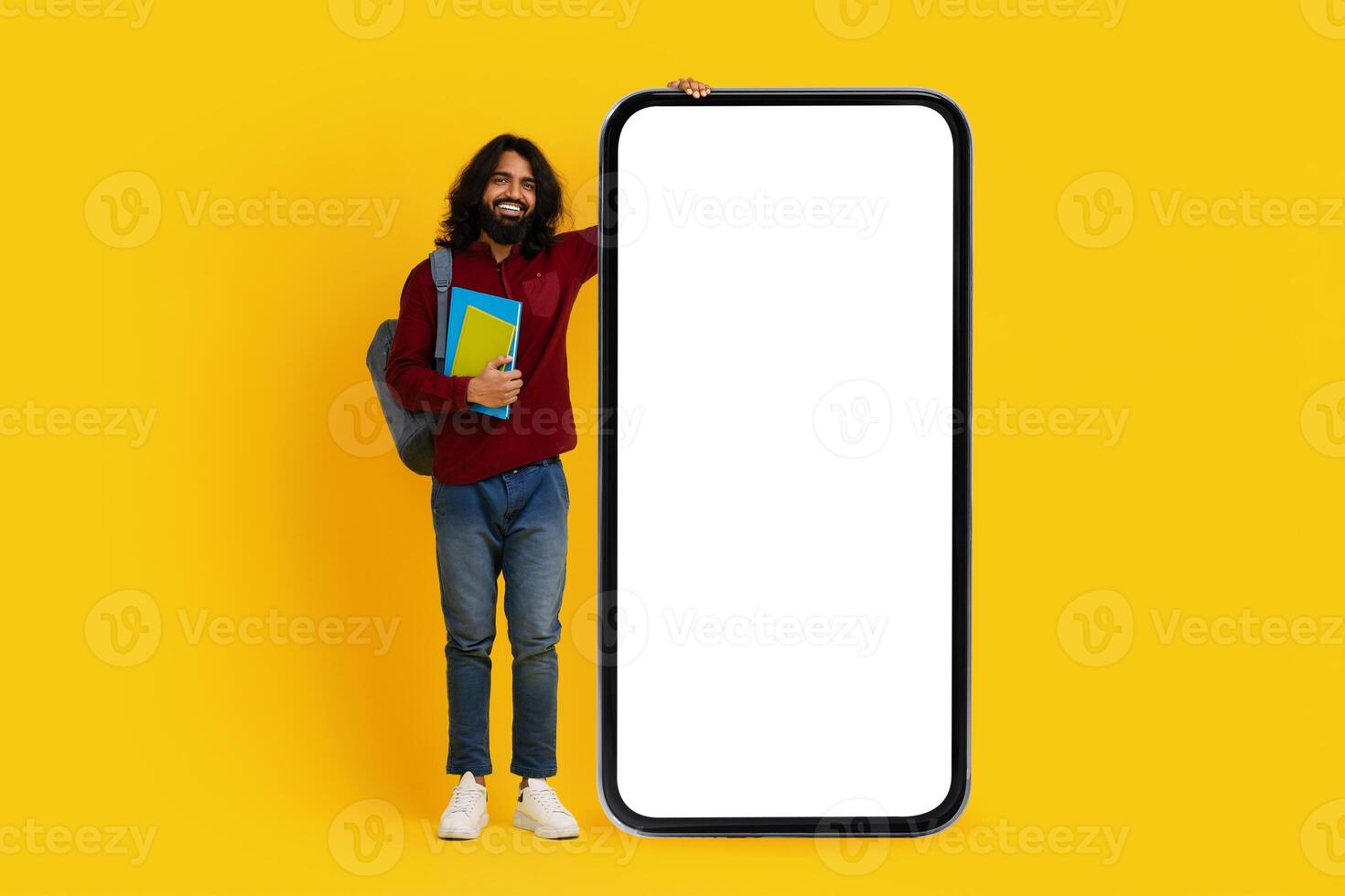 Cheerful Indian man with a beard student stands to the left side, holding colorful books and leaning against a massive, blank screen smartphone photo