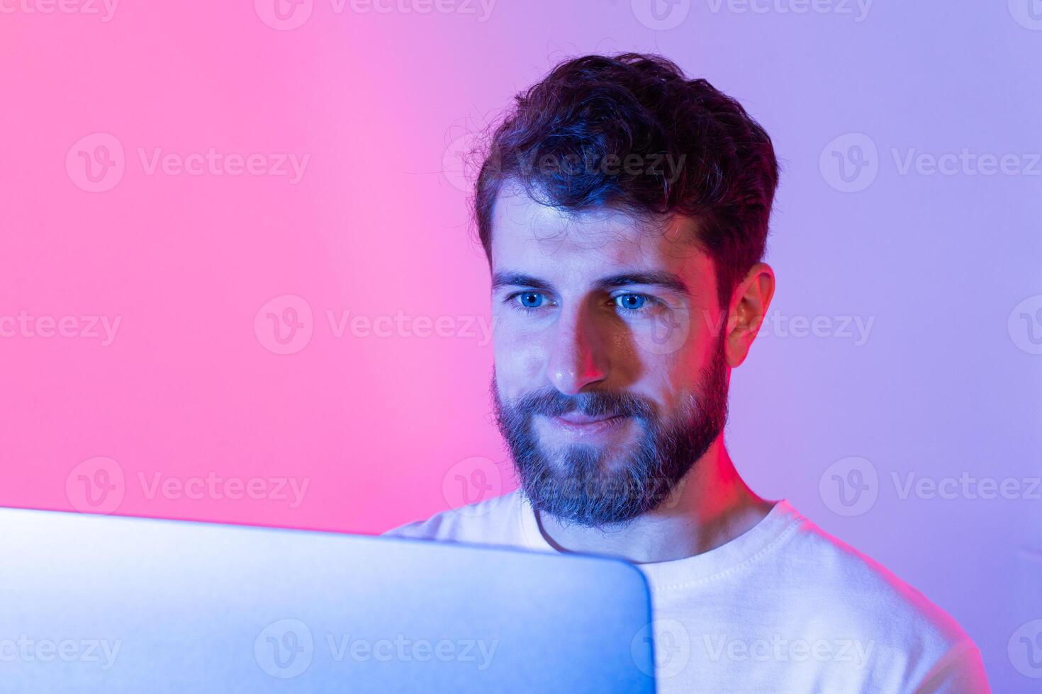 A man with a beard sits in front of a laptop computer, typing and browsing the internet. He is focused on the screen, working or communicating online. photo