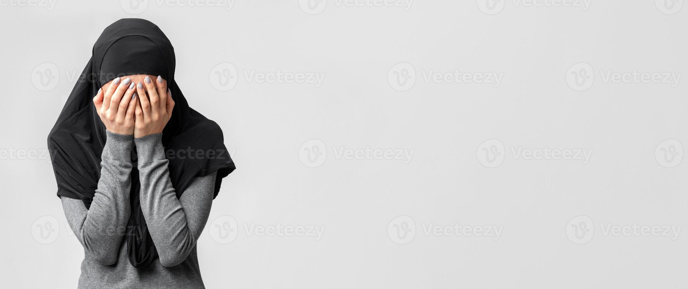 Islamophobia and muslim women discrimination concept. Woman in black hijab covering face with hands, posing over gray studio background photo