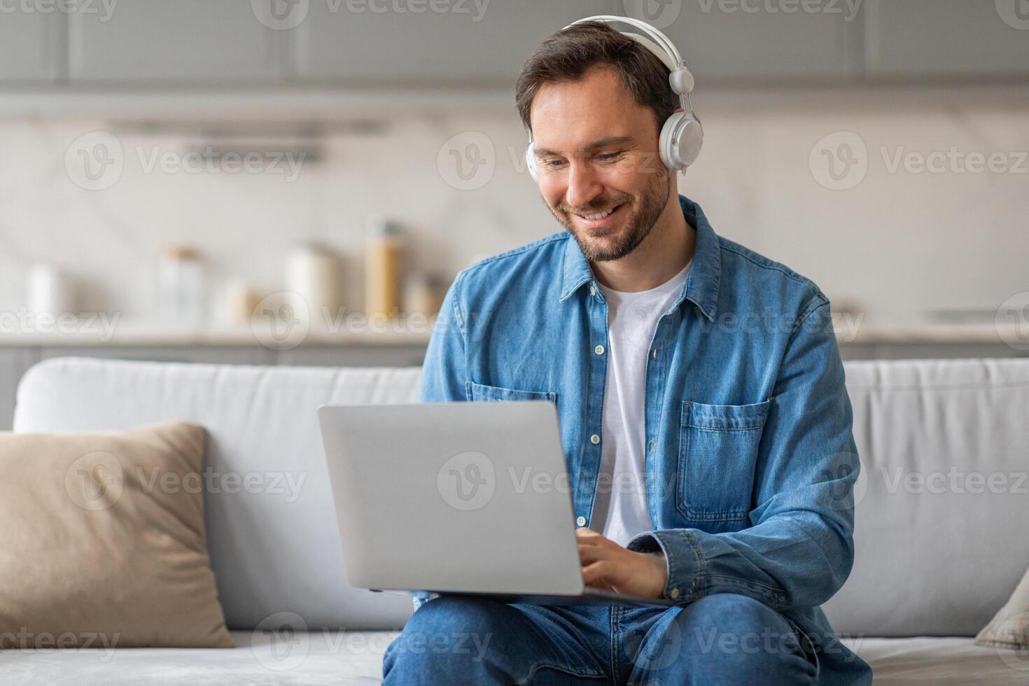 A man in a denim shirt sits on a couch in a living room. He is using a laptop computer and smiling. He is wearing headphones and is focused on his work. photo