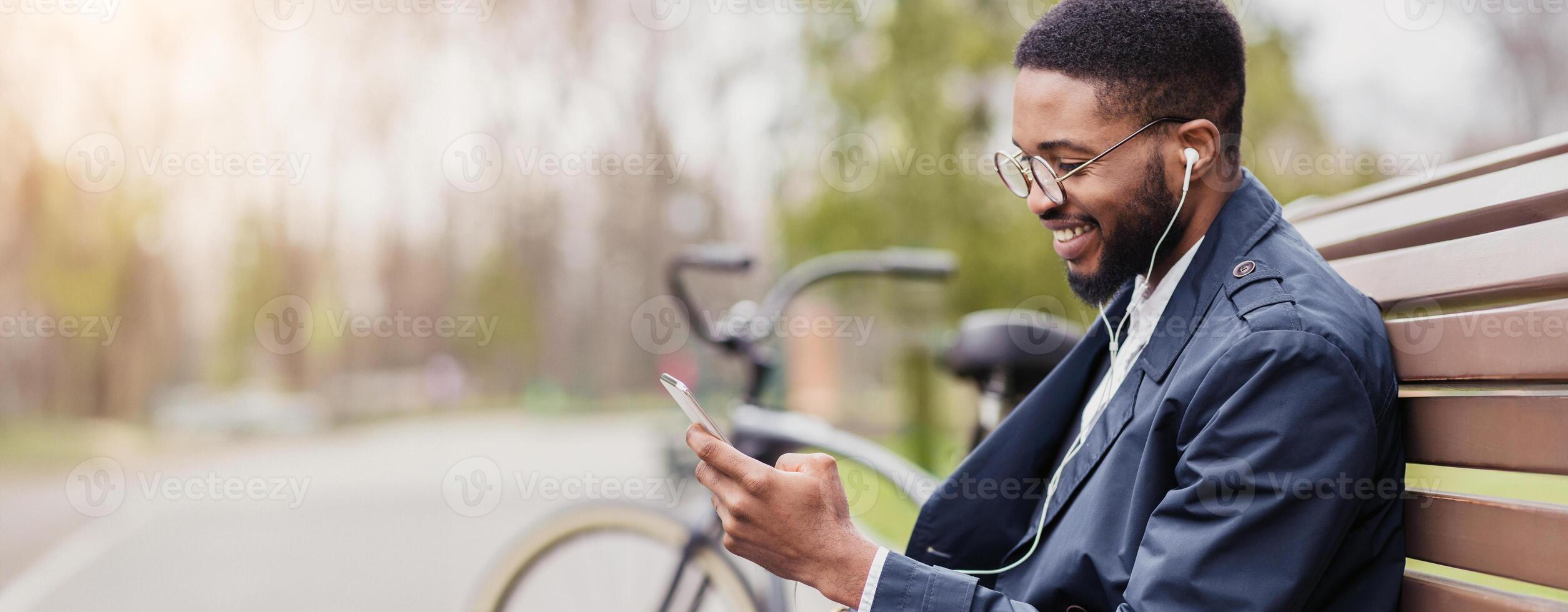 Black business professional is on his phone, sitting on a park bench with a bicycle next to him. The scene is calm and modern, blending work and leisure in a natural setting. photo