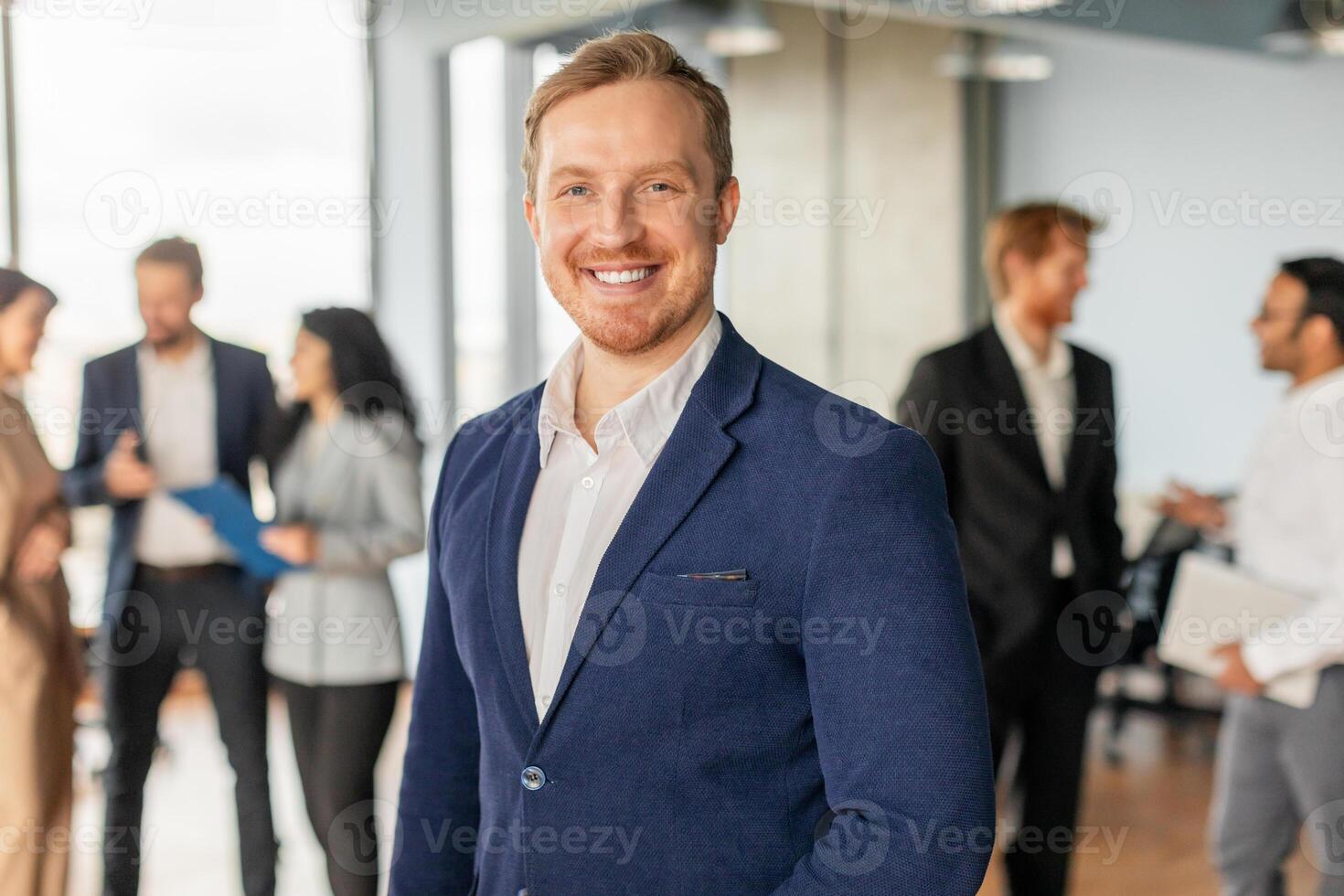 A man in a blue suit and white shirt stands in an office setting, smiling confidently. He is surrounded by colleagues, who are talking and working in the background. The office is bright and modern photo