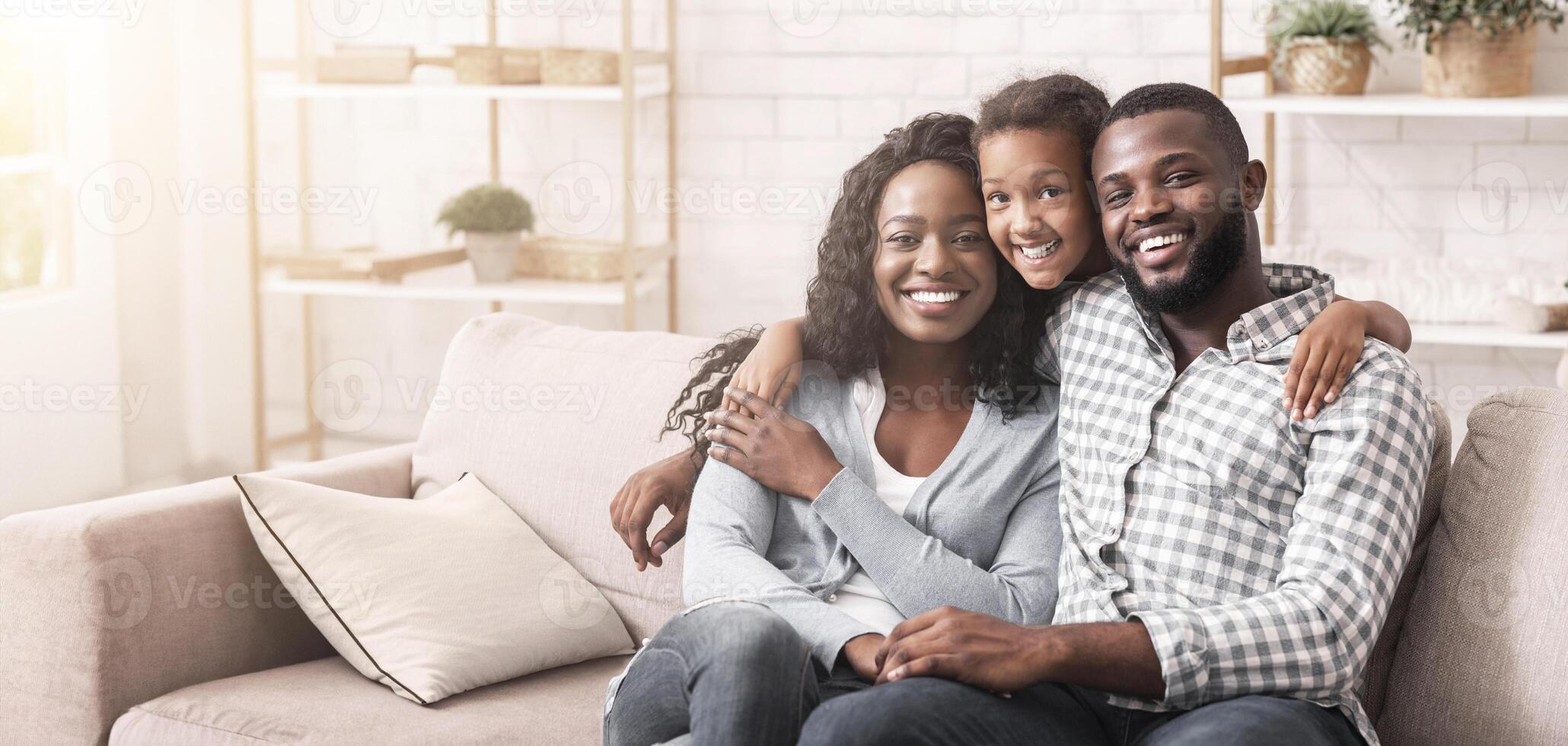 Family portrait. Happy african american parents and their daughter embracing and posing to camera while sitting together on couch at home photo
