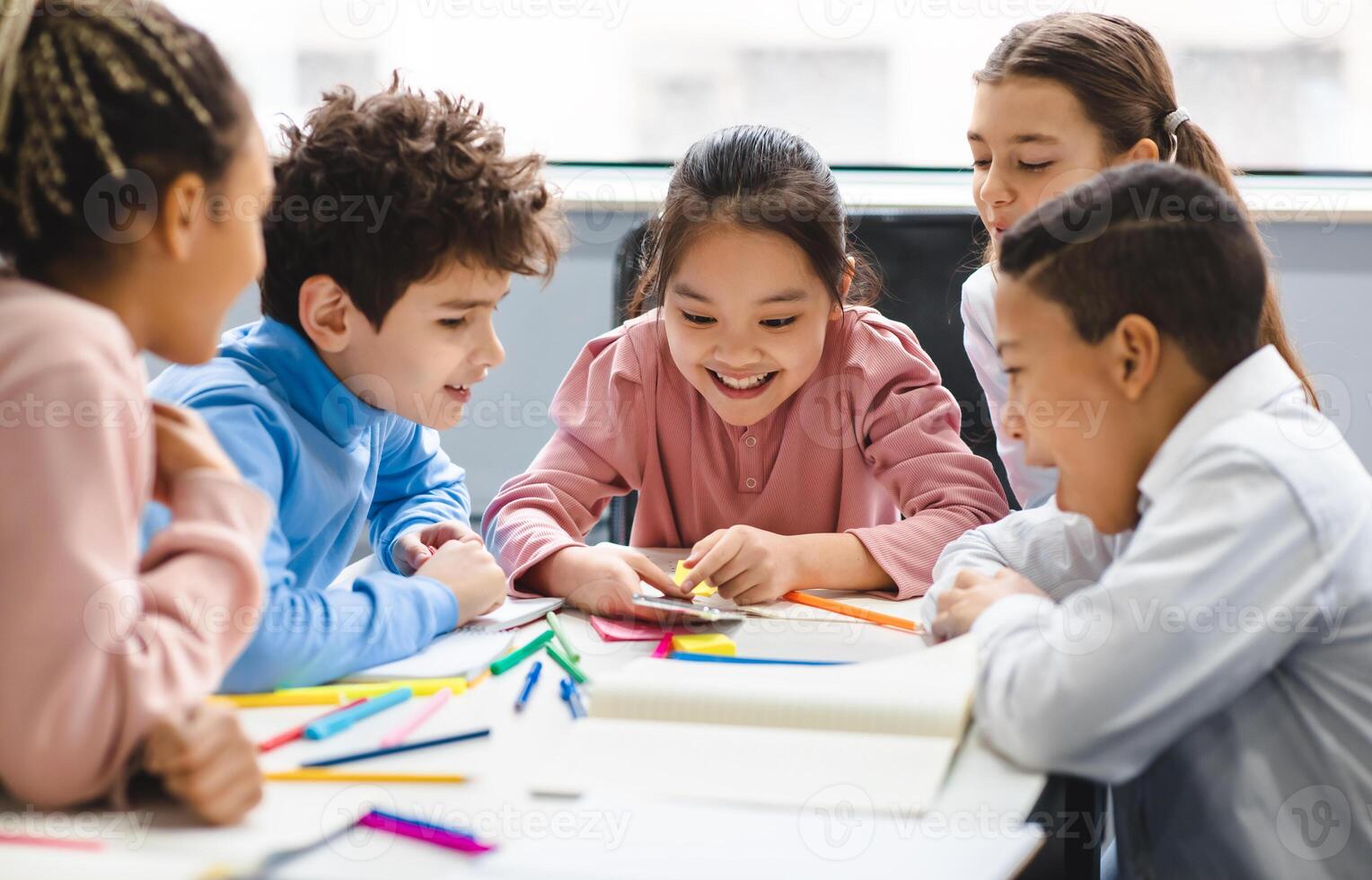 Technology And Pupils Concept. Group of excited multicultural happy junior children sitting at table and using smartphone, playing online mobile games. Modern Device, Gadget Addiction photo