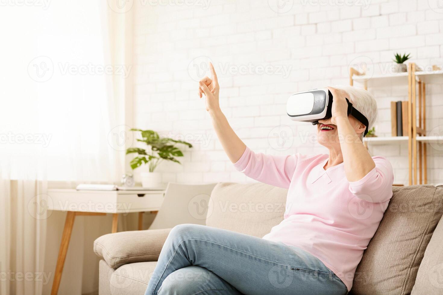 Elderly woman is seated on a couch, wearing a virtual reality headset, engaged in the virtual world, with a hand reaching out in front of her. The room is dimly lit, with the headset glowing blue. photo