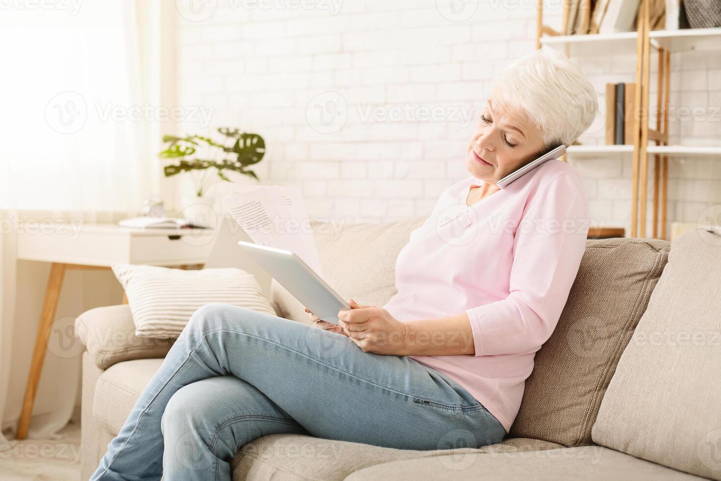 Busy senior woman consulting client on phone and reading contract on digital tablet at home, empty space photo