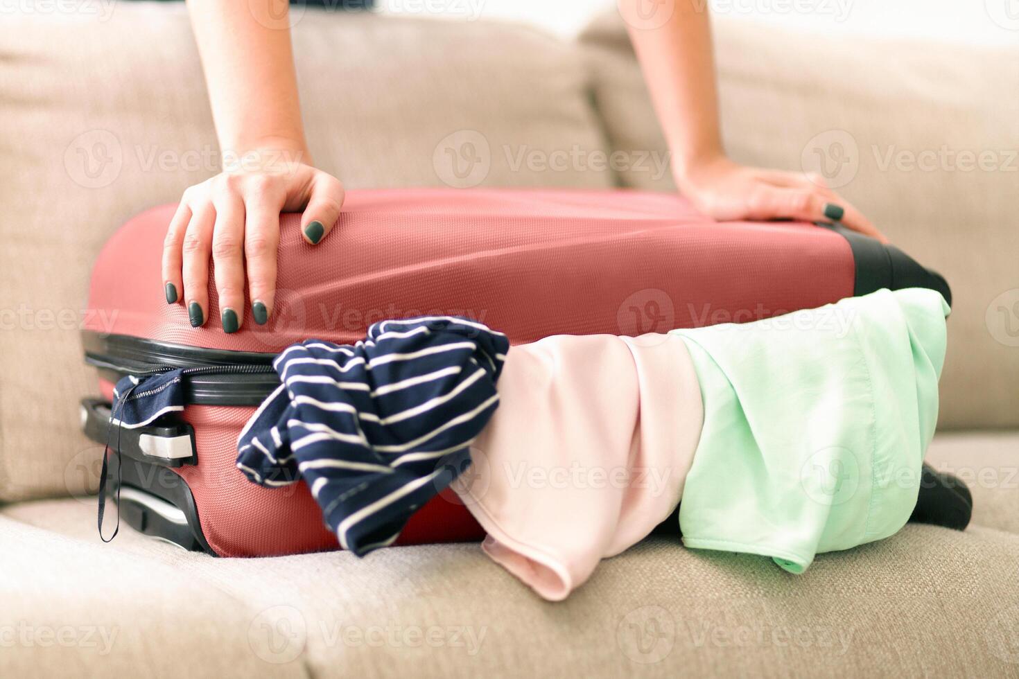 Cropped of woman trying to close an overstuffed luggage compartment on what appears to be a sunny day. Various clothes are spilling out from the sides, packing for a new journey. photo