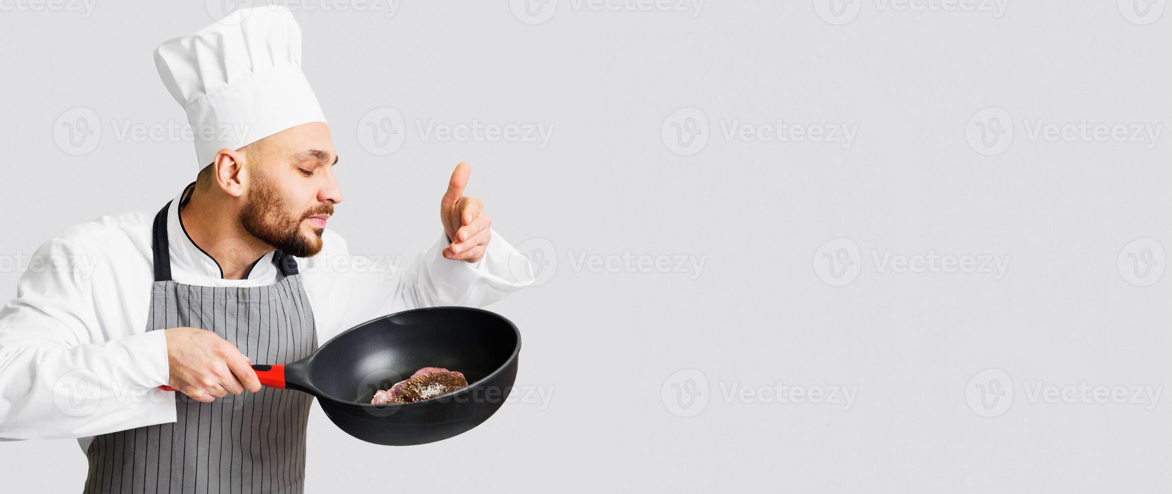 Male Chef Cooking Meat Smelling Frying Pan With Fresh Beef Steak Standing Over Gray Studio Background. Panorama photo