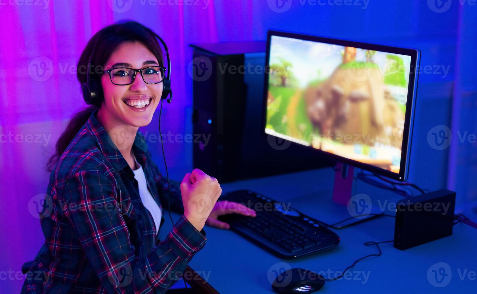 A young woman sits in front of a computer screen, wearing a headset and smiling as she plays a game. The computer monitor is illuminated by neon pink and blue lighting photo