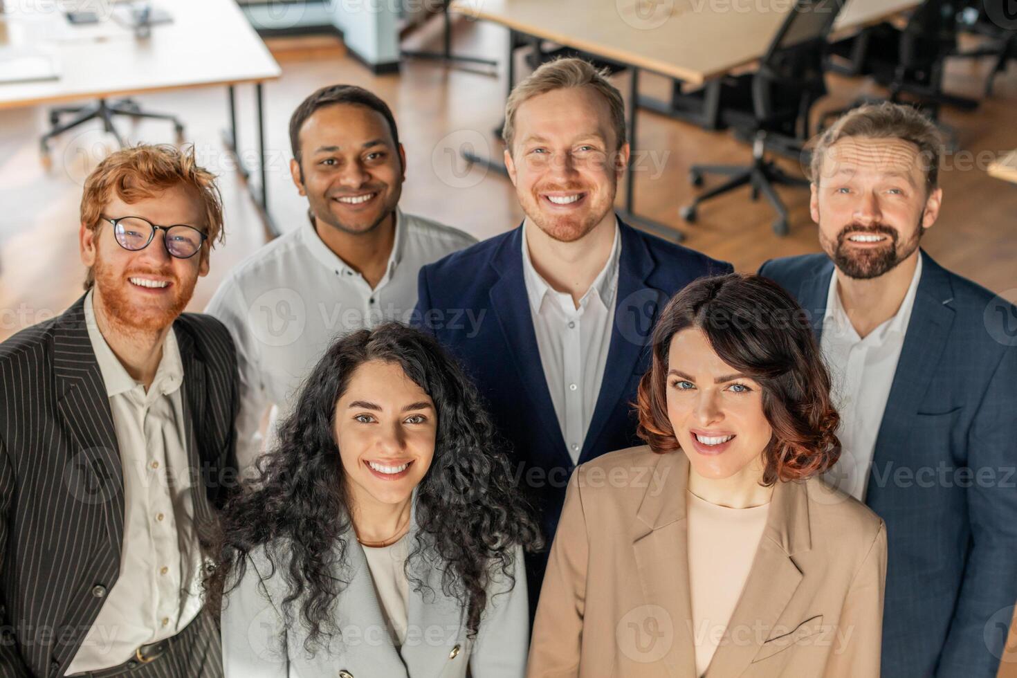 Five diverse business professionals stand together in a modern office, looking directly at the camera and smiling. They are wearing business casual attire, high angle view photo