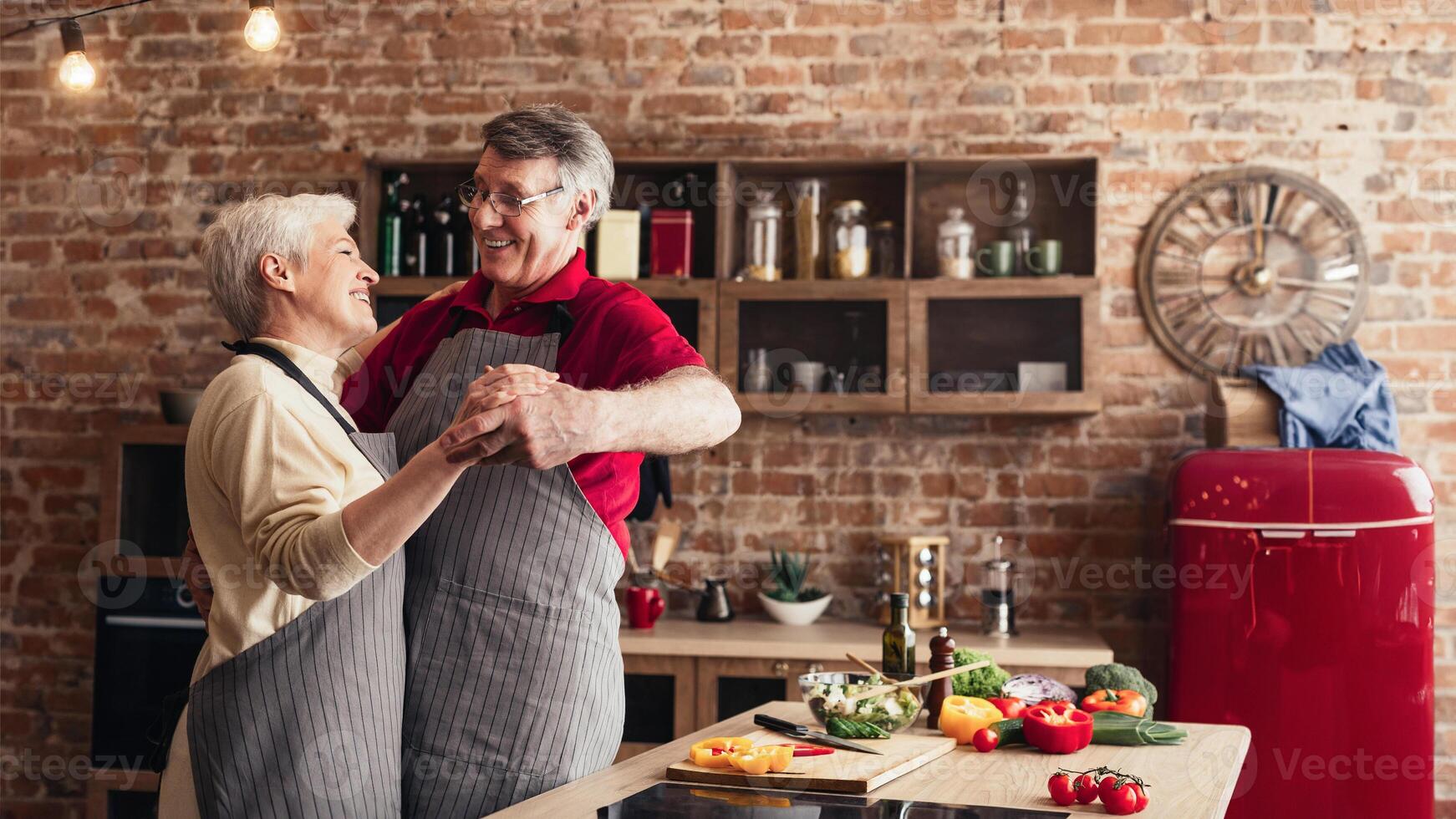 A senior couple in aprons, smiling and holding each other, are dancing in a kitchen. Fresh vegetables are on the counter in front of them. photo