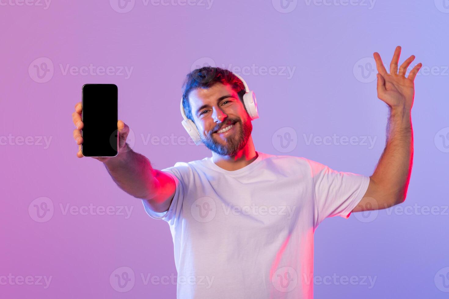 A man wearing headphones holds up a cell phone, with a focused expression on his face. He appears to be listening to music or talking on the phone. photo