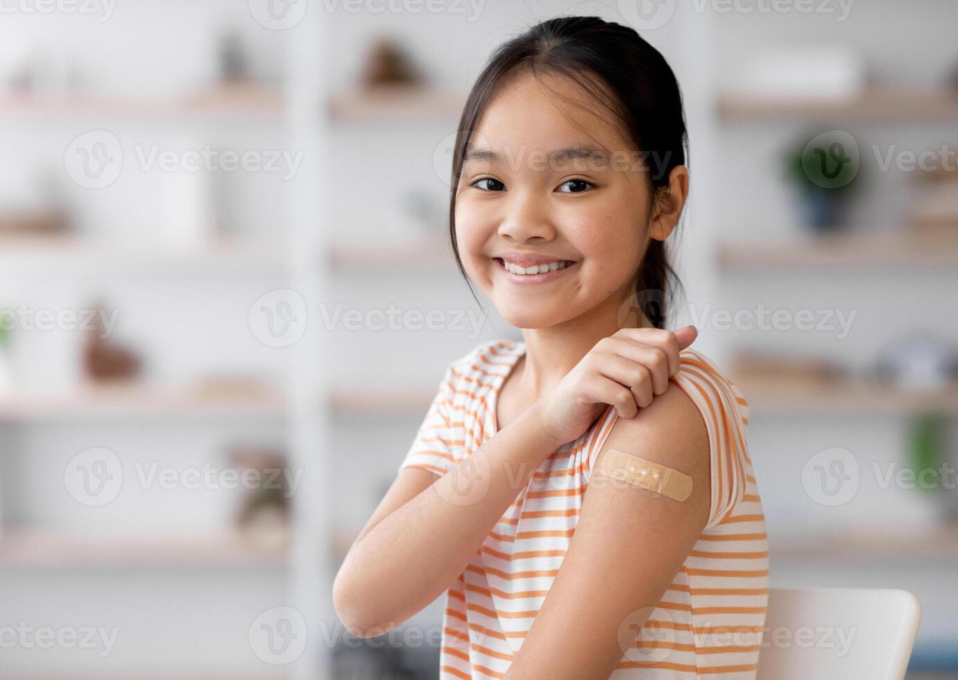 Healthy asian kid girl got vaccinated, sitting on couch at home and showing adhesive band on her shoulder, smiling at camera, panorama with copy space. Kids vaccination, immunization concept photo