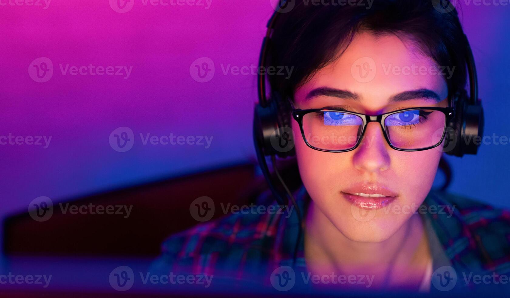 A woman wearing glasses and headphones is looking intently at a computer screen. The room is lit in a purple and blue color scheme, creating a vibrant and modern atmosphere photo