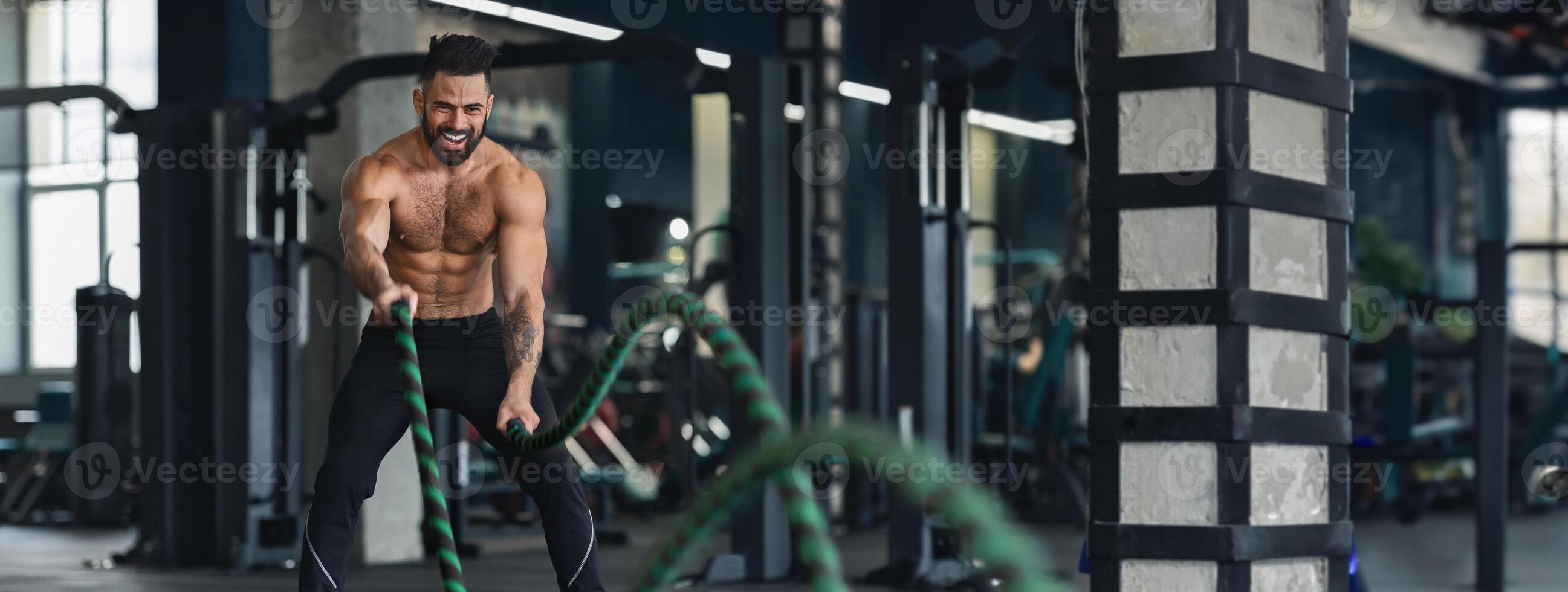 Crossfit training. Naked muscular man exercising with battle ropes at gym, free space, panorama photo