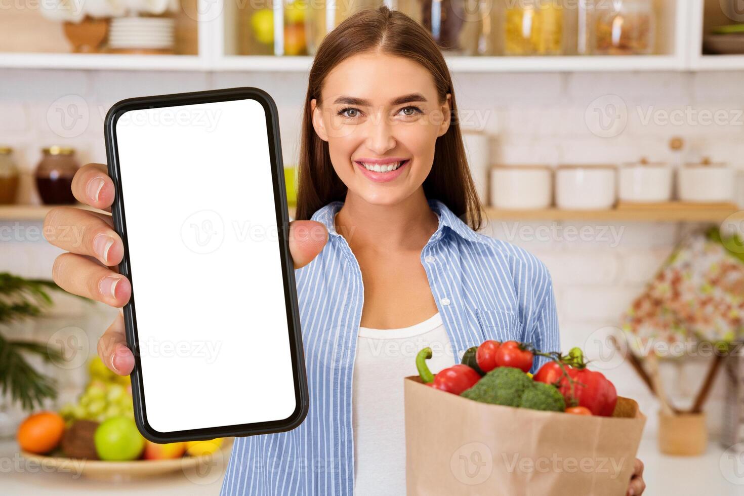 A woman in a kitchen holds a brown paper bag filled with fresh vegetables while showing a smartphone with a blank screen. photo