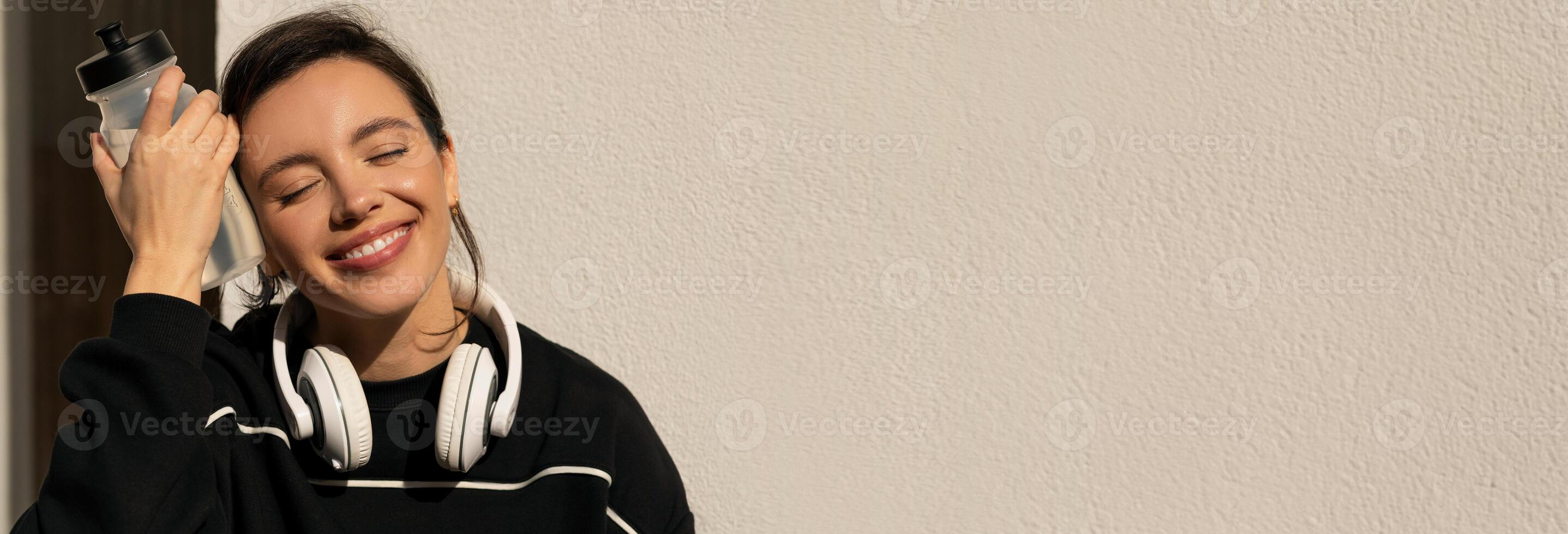 Smiling caucasian millennial lady in sportswear and headphones relaxing with bottle of water on street, enjoy break, close up. Sports ad and offer, body care, weight loss and rest, copy space photo