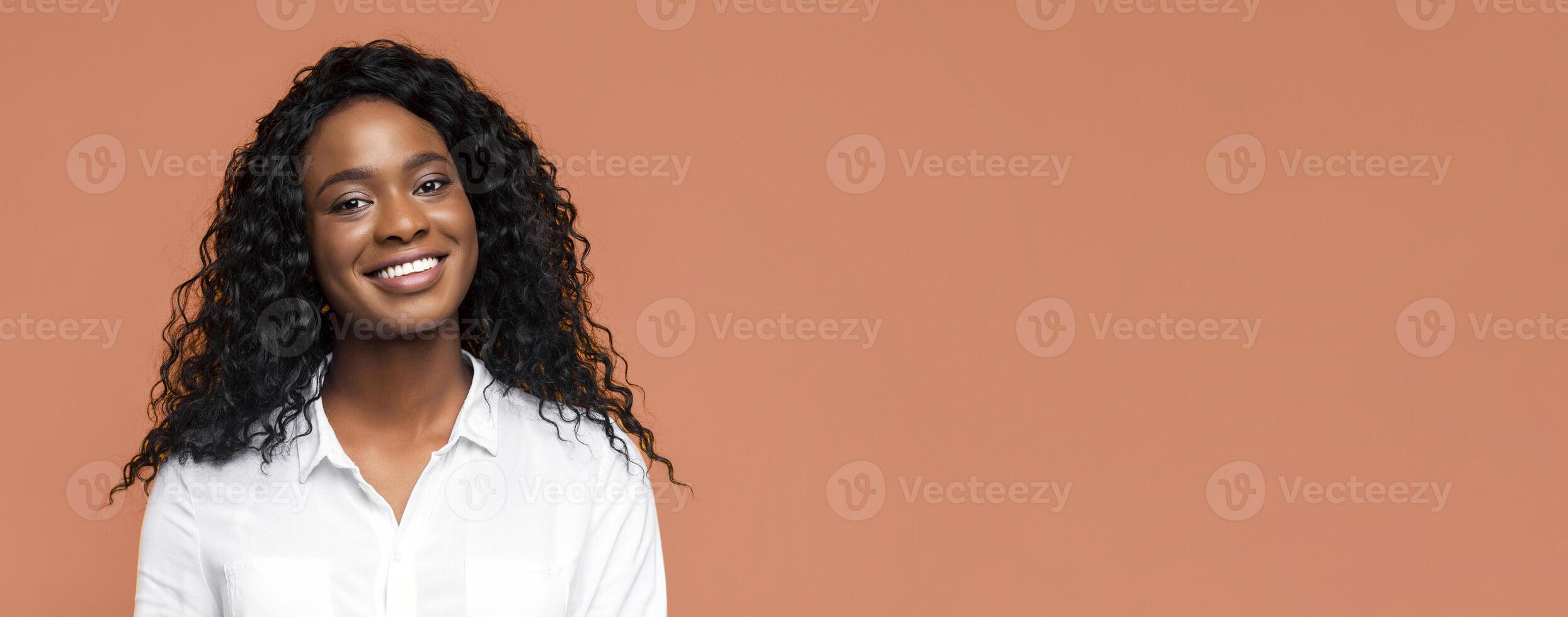 Black woman smiling warmly at the camera. She is wearing a white shirt and her dark curly hair is pulled back from her face. The background is a simple tan color photo