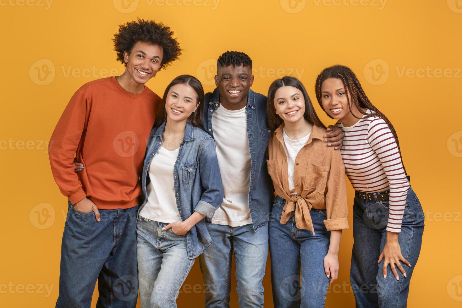 Group of five diverse young adults standing in front of a yellow background. They are all smiling and looking at the camera. The group appears to be friends and is enjoying each others company. photo