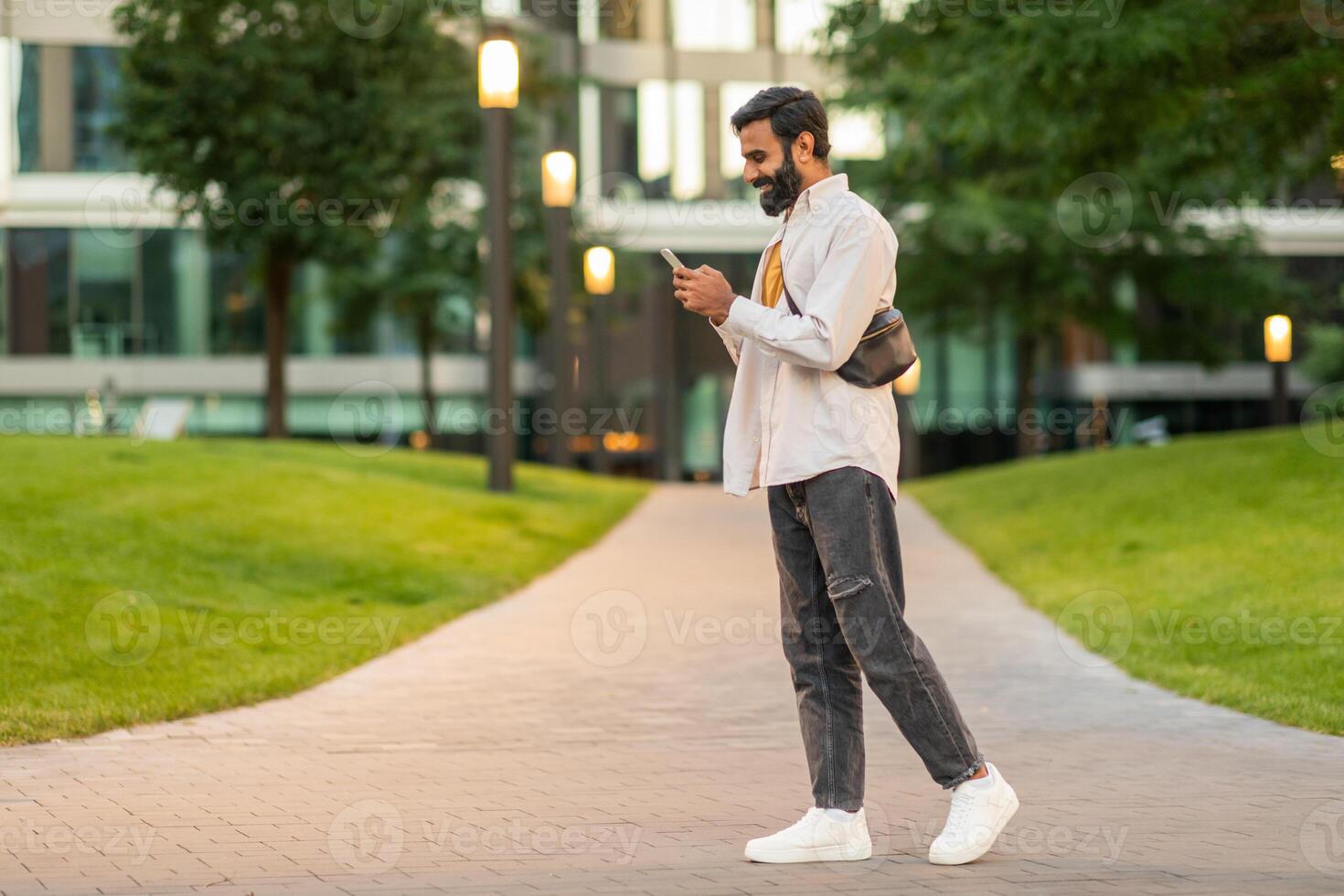Indian man is walking down a sidewalk while engrossed in his cell phone, his attention focused on the screen rather than his surroundings, multitasking as he navigates the urban environment. photo