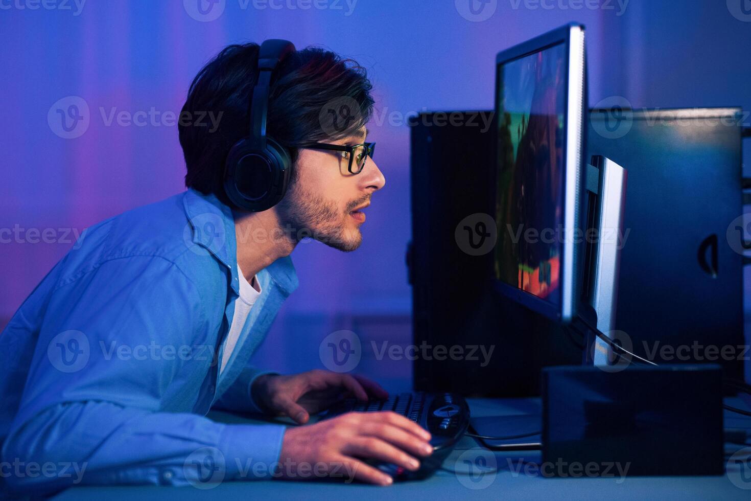 A young man with headphones and glasses is intensely focused on his computer screen, playing a game. The room is dimly lit with blue and purple hues, creating a mood of late-night gaming photo