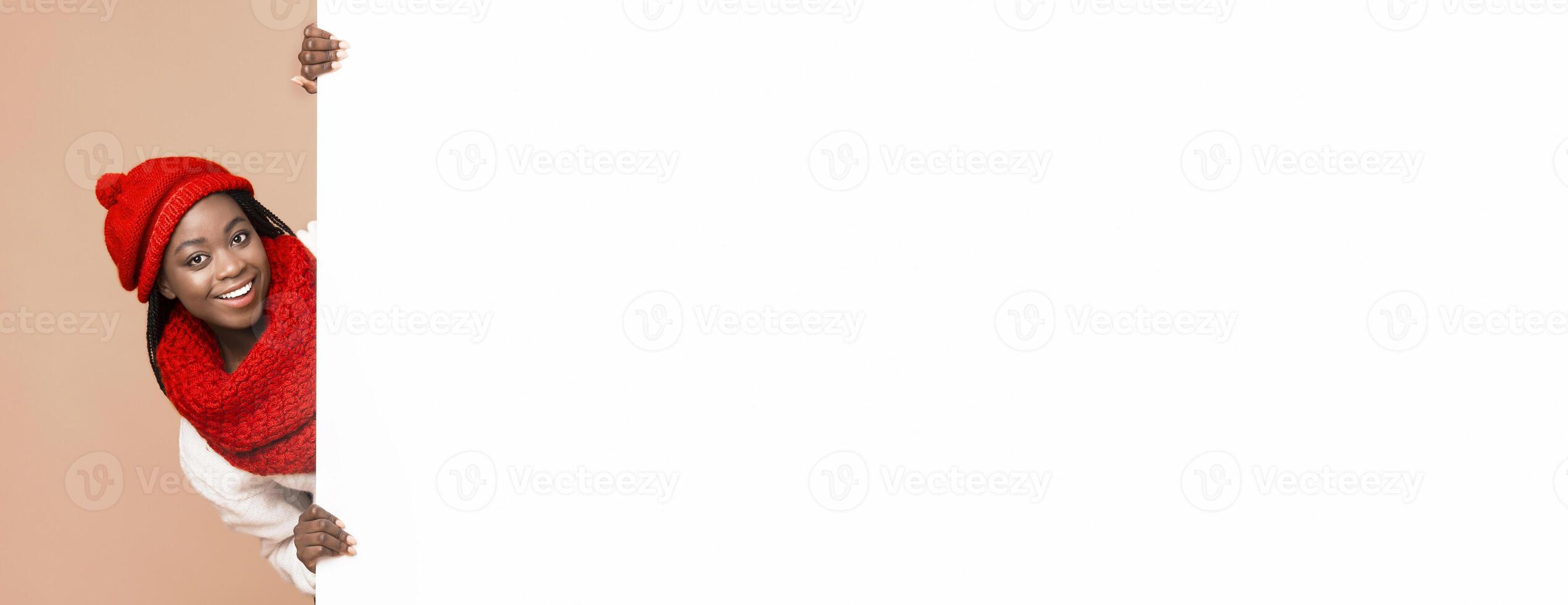 Empty Space For Advertising. Black cheerful woman in red knitted set staying next to empty beige board, beige background photo
