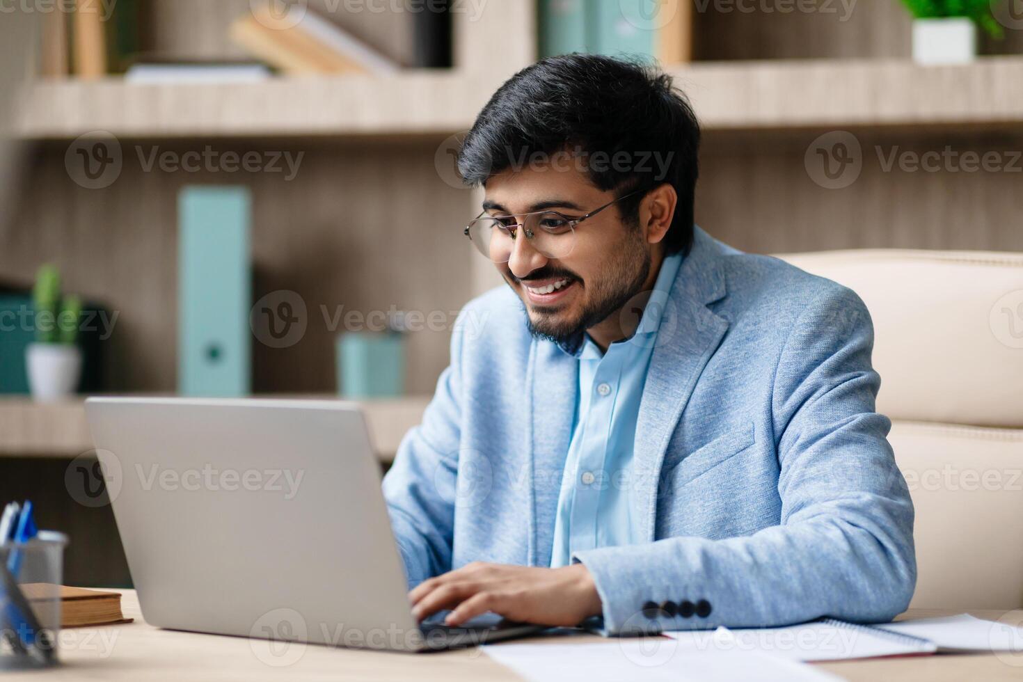 Indian man wearing a light blue jacket sits at a desk in a modern office, smiling as he works on his laptop computer. He appears to be in a positive and focused mood. photo