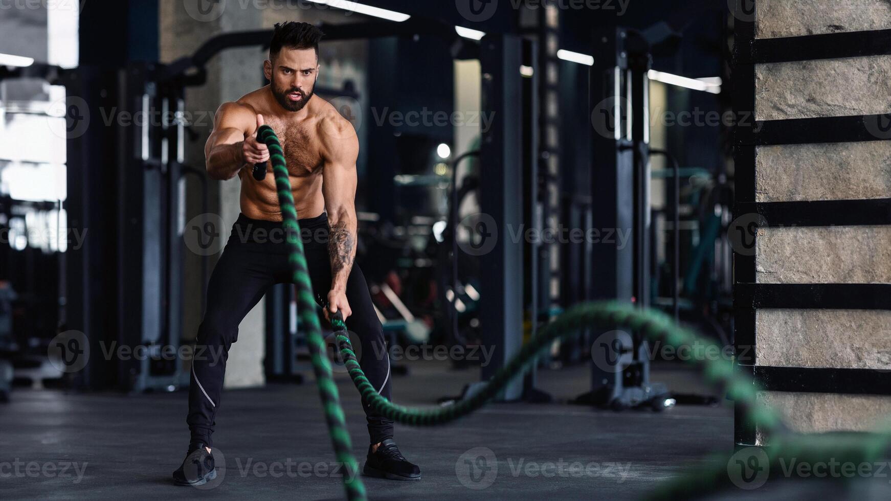 A muscular man is working out with battle ropes in a gym. He is wearing black shorts and shoes. He is focused on his workout. photo