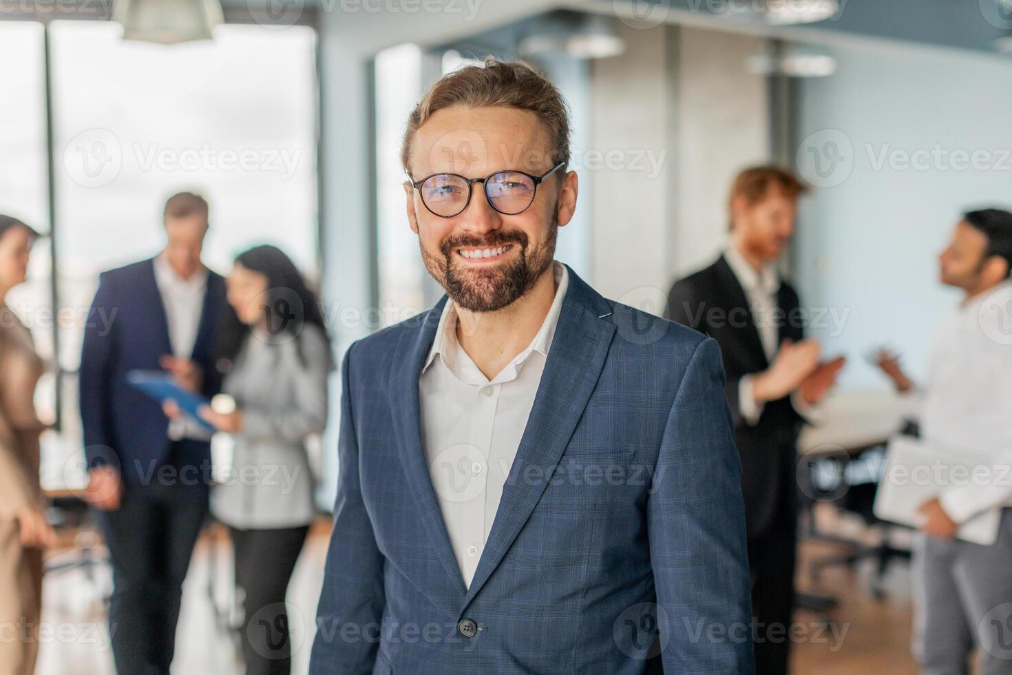 A businessman wearing a blue suit and glasses smiles while standing in a modern office setting. Other office workers are standing in the background and engaged in conversations. photo