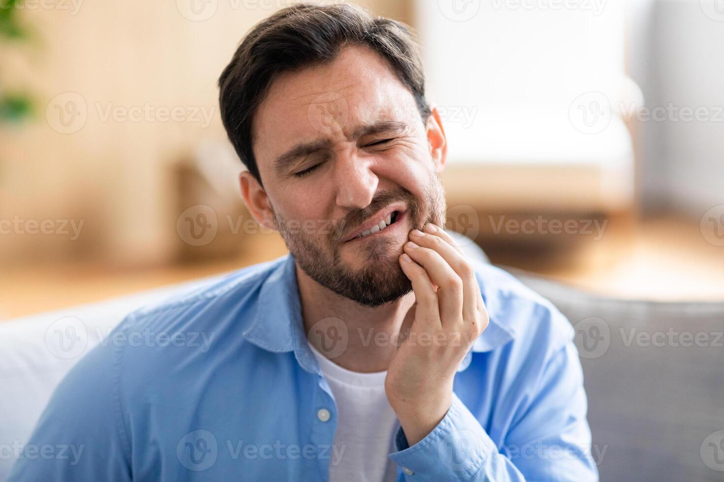 A man is shown sitting on a couch, visibly in discomfort, holding his jaw with a pained expression due to a toothache. He appears to be seeking relief or contemplating seeking dental help. photo