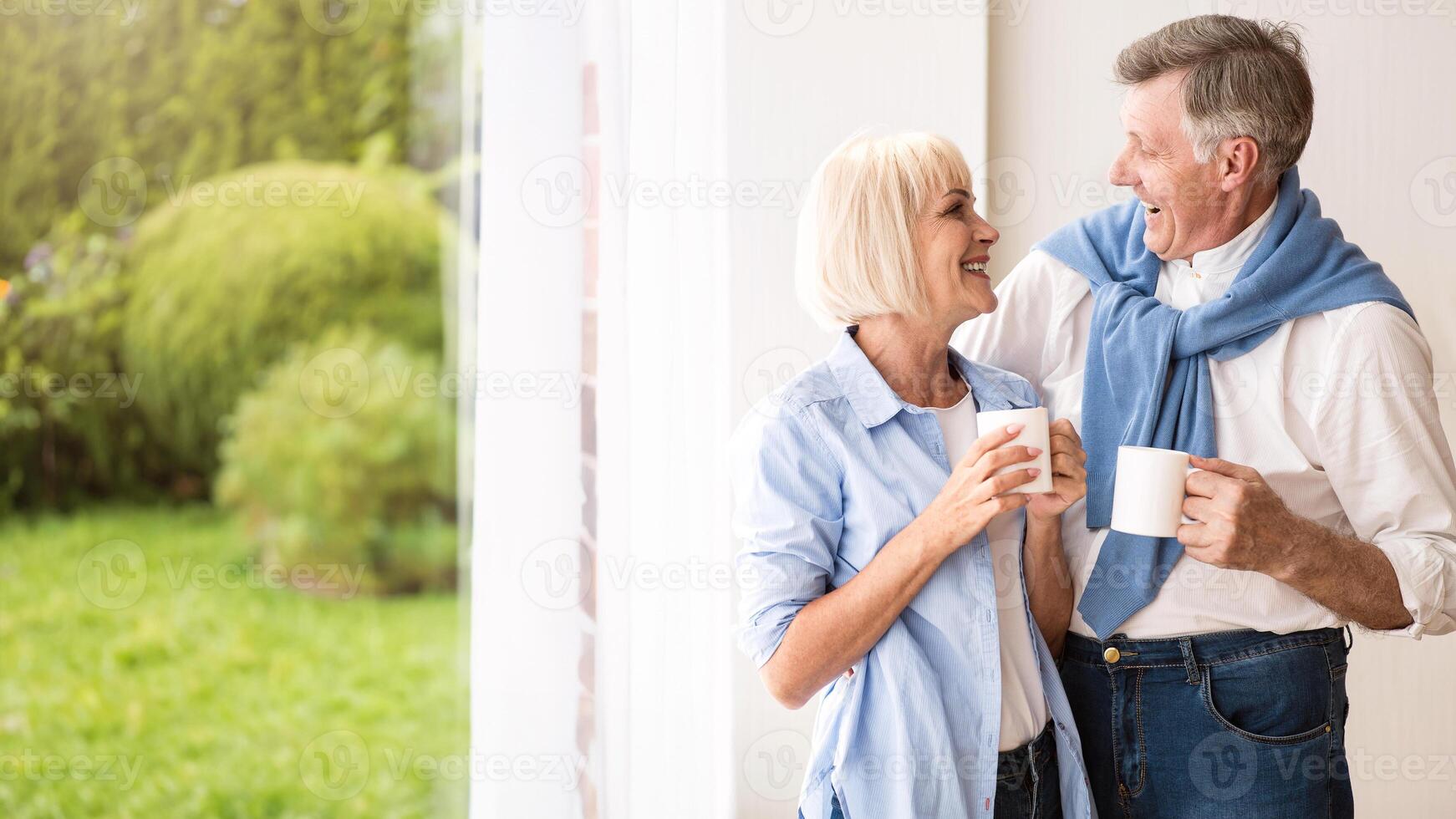 Happy senior couple talking near window and drinking tea, copy space photo
