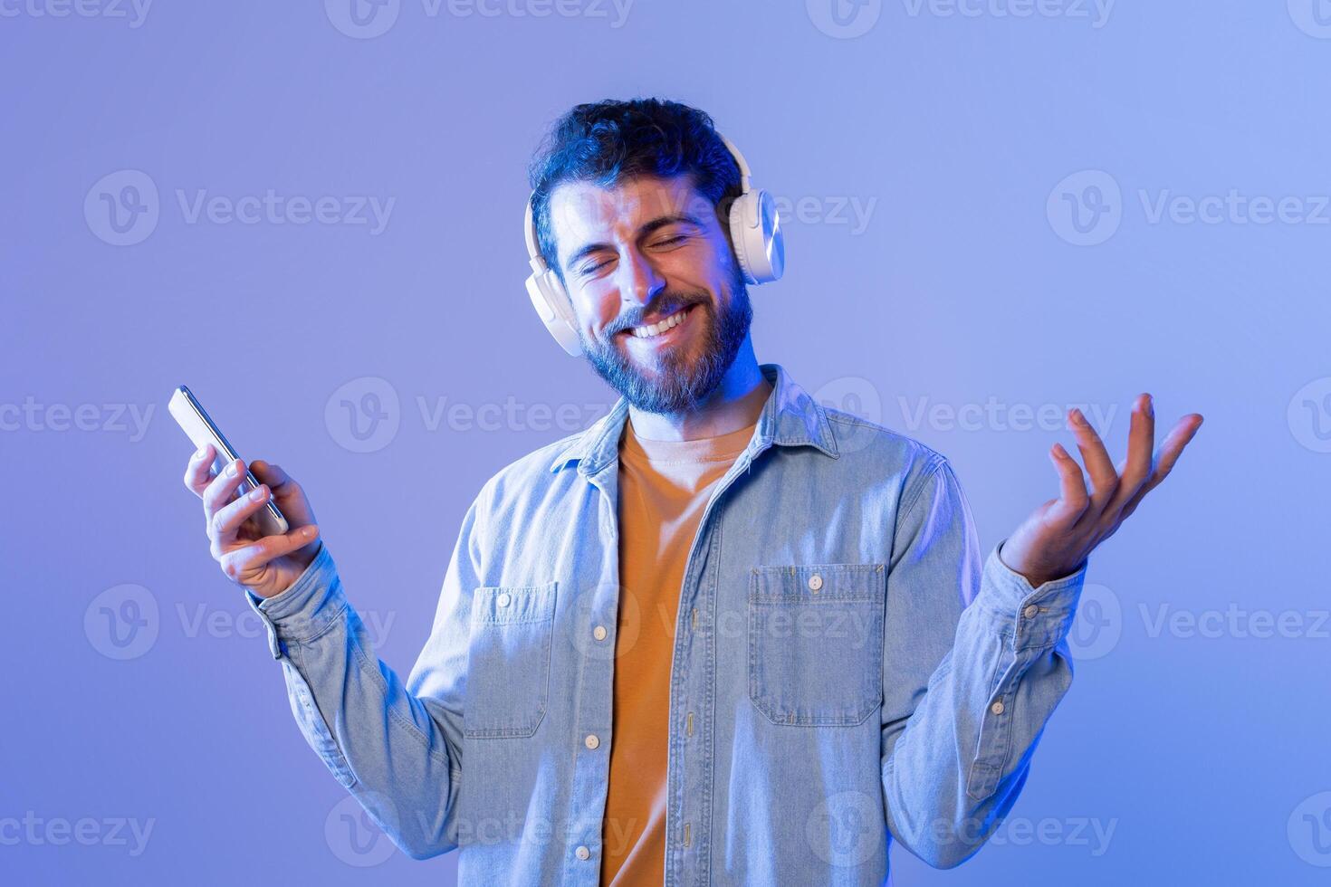 A man with headphones on his ears is seen holding a cell phone in his hand. He seems focused on the screen, possibly listening to music or a phone call. The background is not visible in the frame. photo
