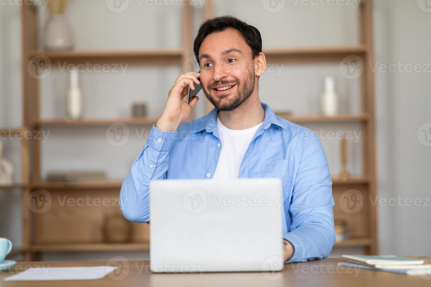 A man sitting at a desk, engaged in a phone conversation. He is multitasking, with a laptop open in front of him. photo