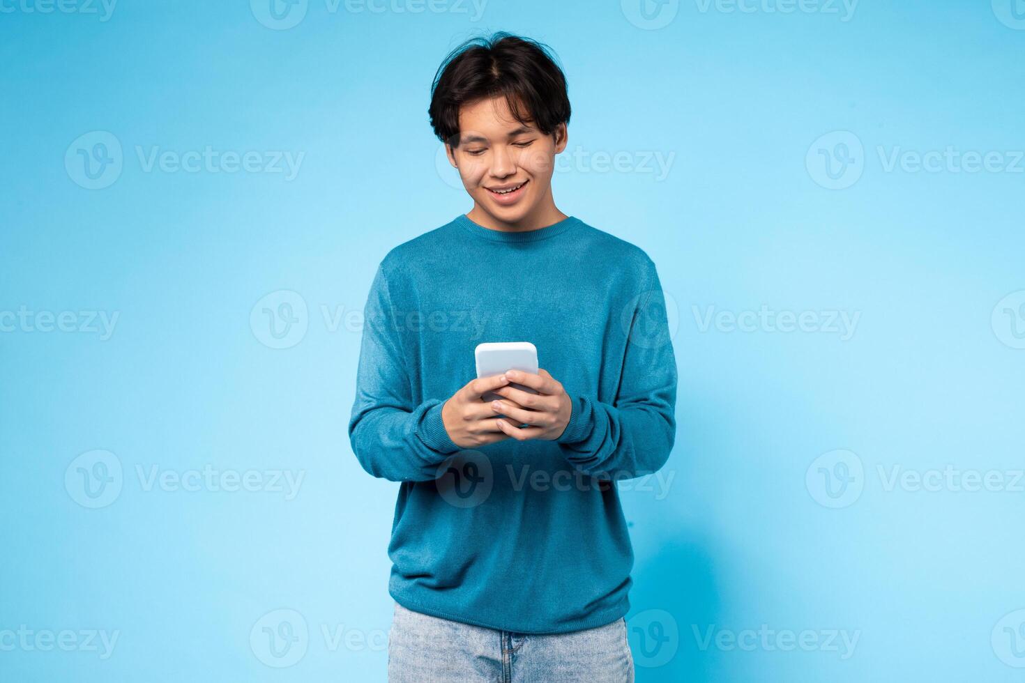 Asian guy wearing a blue sweater is sitting or standing and looking intently at a cell phone in his hands. He appears to be focused on the screen, possibly browsing, reading, or texting on the device. photo