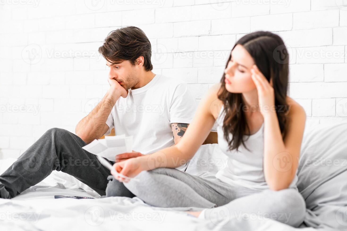 A man and a woman are sitting on a bed, looking distressed. The man is holding a piece of paper in his hand, and the woman is looking down at the floor. They both appear to be troubled or worried. photo