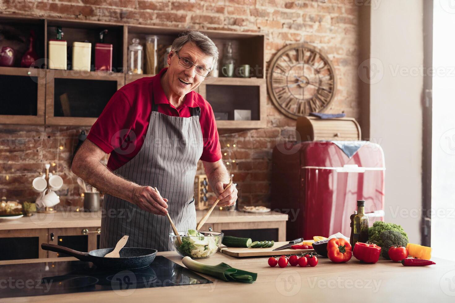 A senior man wearing a grey apron and a red shirt is preparing a salad in his modern kitchen. He is standing at the counter, holding a wooden spoon in each hand, and stirring a bowl of green salad photo