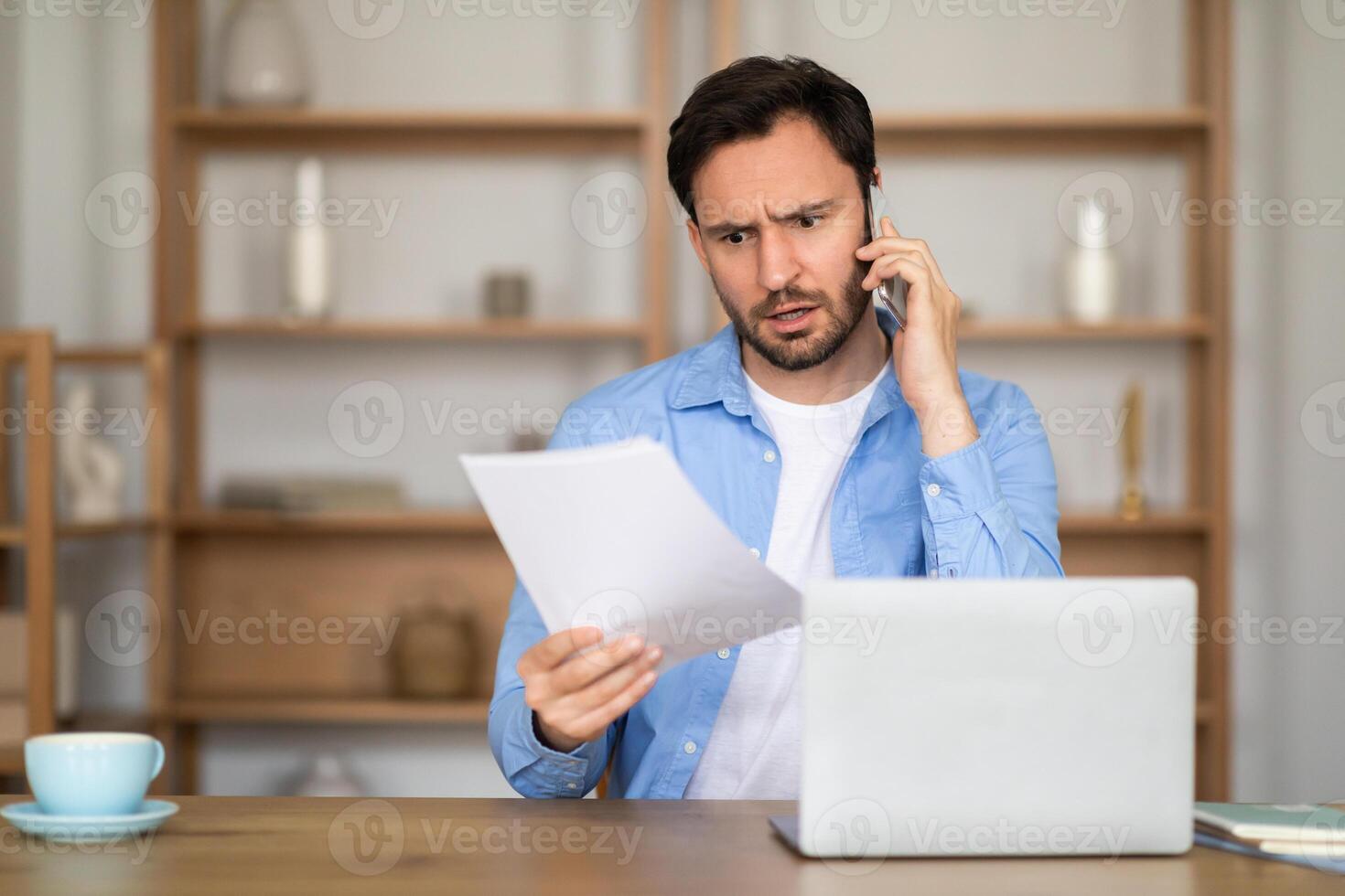A man appears engaged in a serious conversation on his cellphone, simultaneously examining a document with a furrowed brow. The scene takes place in a well-lit home office photo