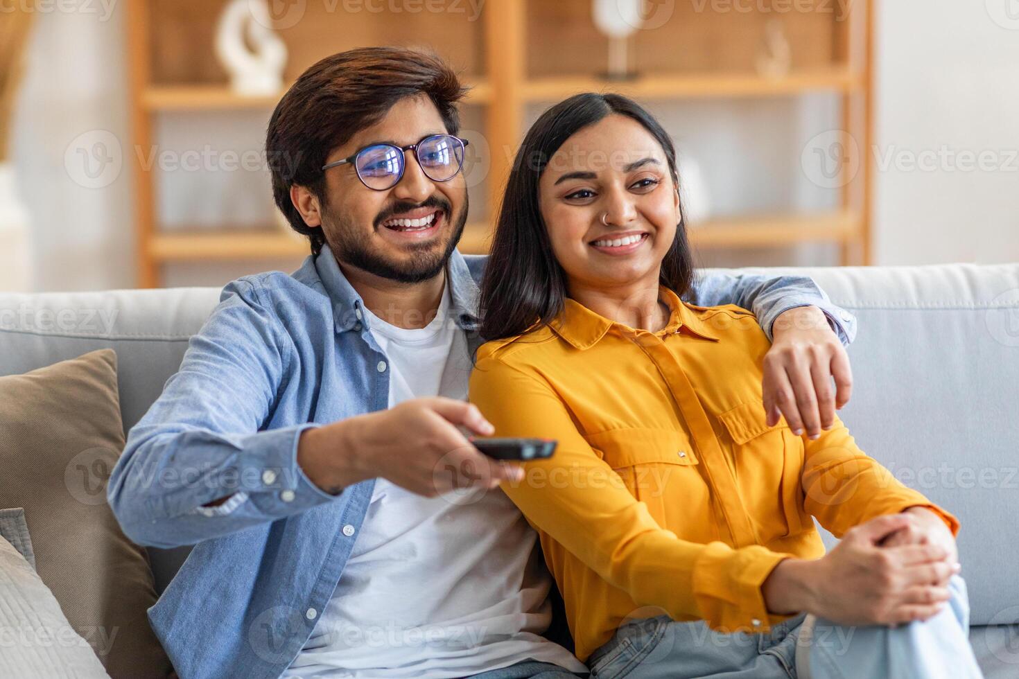Indian man and woman are seated on a couch, holding a remote control. They appear to be watching television together, immersed in the content being displayed on the screen. photo