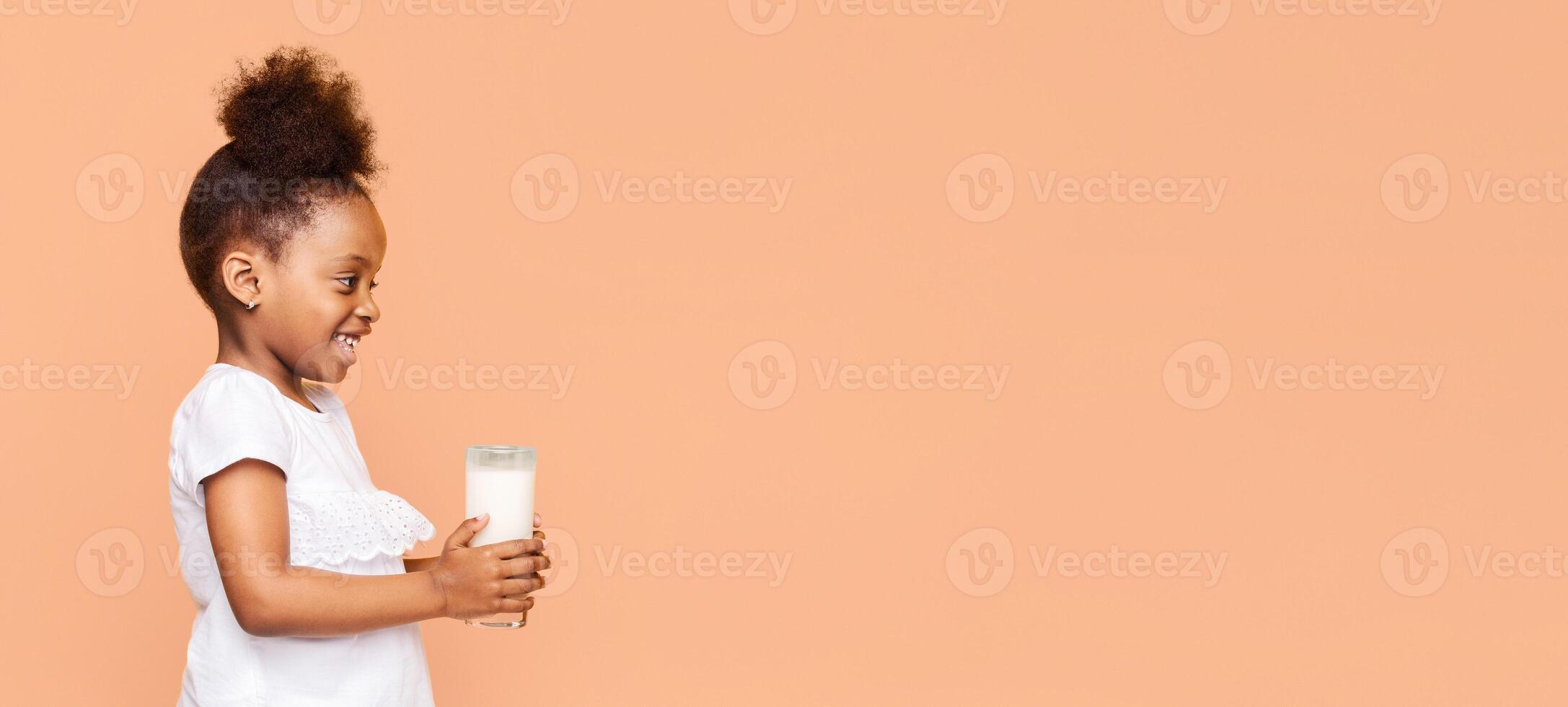 A little back girl with curly hair smiles as she holds a glass of milk in front of a peach-colored background. She is wearing a white shirt with ruffles and looks happy and healthy, copy space photo