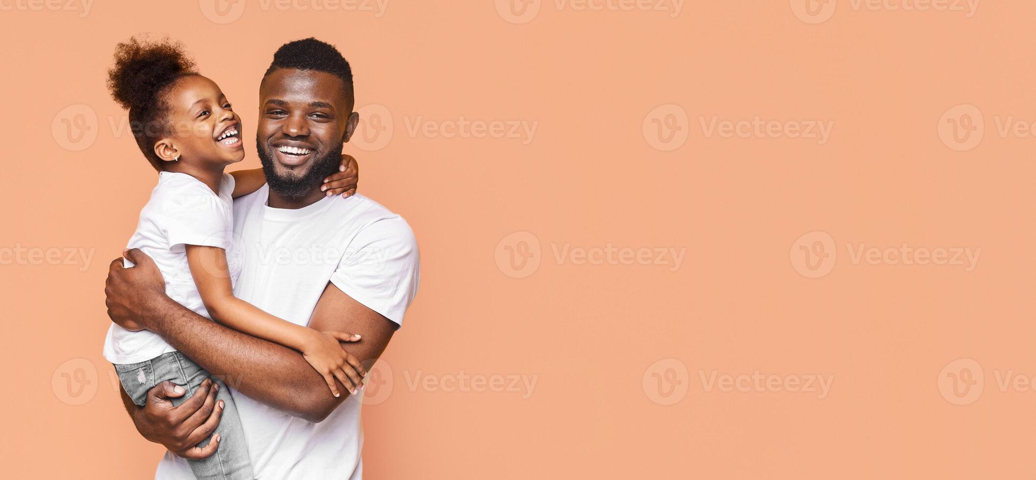 A smiling black father holds his daughter in his arms while looking at the camera. The daughter is also smiling and looking at the camera, copy space photo