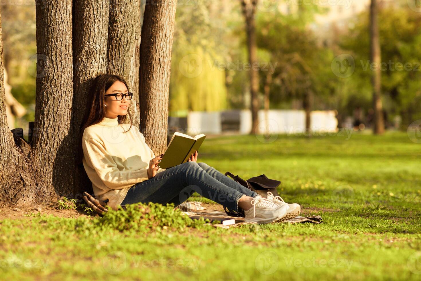 Korean woman is sitting under a tree, engrossed in a book she is reading. The tree provides shade as she enjoys her quiet reading time outdoors. photo