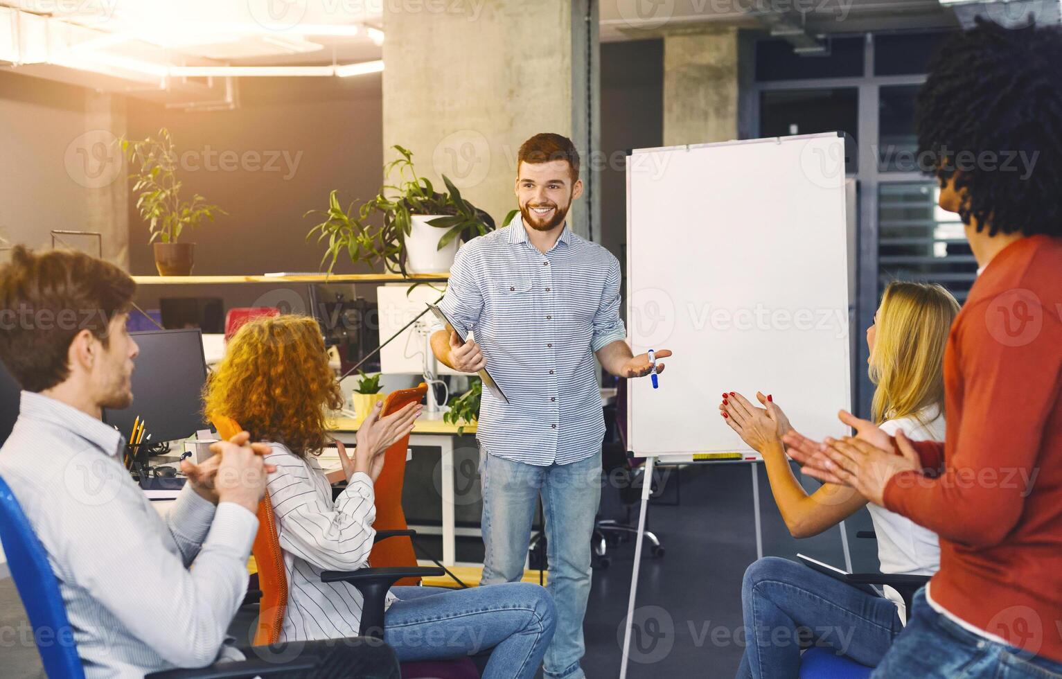 A man stands in front of a group of attentive individuals, gesturing and speaking confidently as he delivers a presentation. The audience is seated, listening intently, taking notes photo