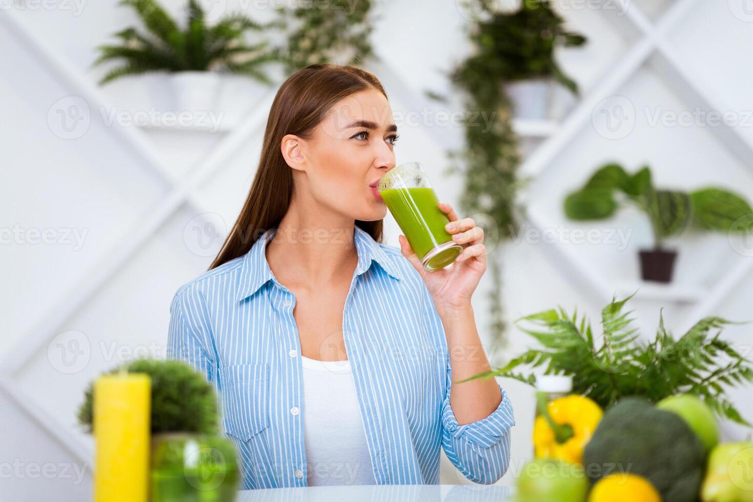 A woman in a blue shirt sits at a table in a kitchen and drinks a green smoothie. Surrounding her are fruits and vegetables, and there are plants in the background. photo
