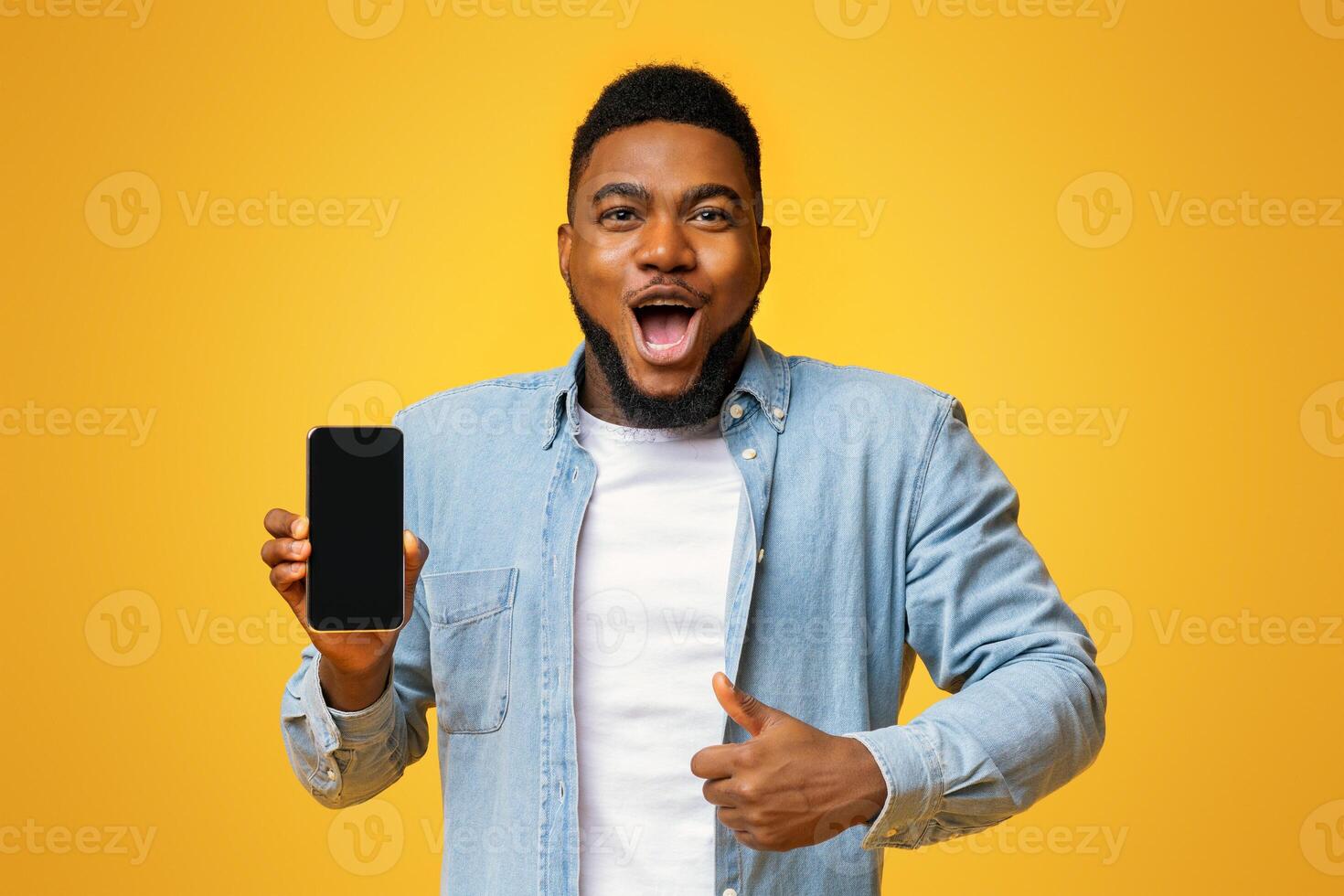 Wow, amazing app. Surprised african man holding smartphone with black screen and showing thumb up, yellow background with free space photo
